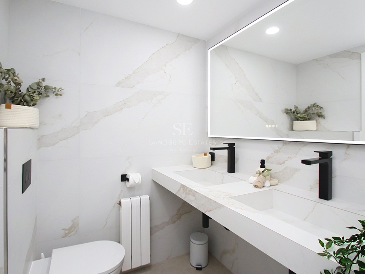 Baño moderno con paredes efecto mármol blanco, lavabo doble con grifería negra mate y gran espejo retroiluminado.