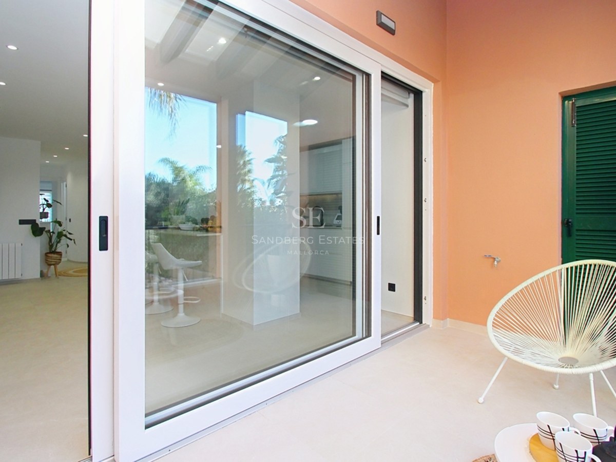 Terraza luminosa con silla Acapulco blanca y puertas correderas de cristal que abren hacia un salón moderno.