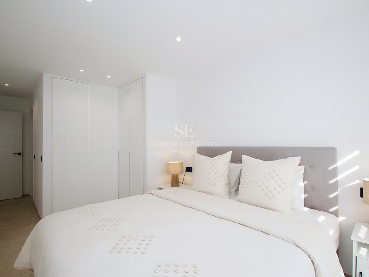 Un dormitorio moderno y luminoso con ropa de cama blanca, cabecero gris, armarios integrados y luz natural.