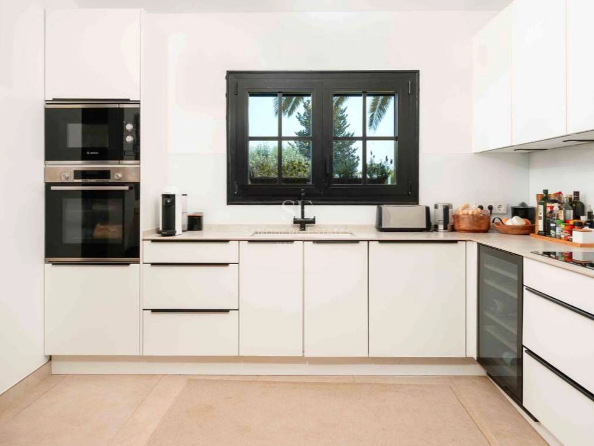 Contemporary white kitchen featuring a black-framed window, integrated oven, wine cooler, and minimalist stone countertops.
