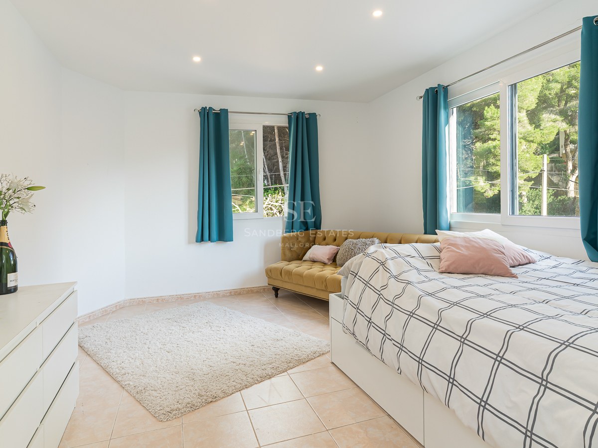 Bright bedroom with a bed, mustard yellow velvet sofa, teal curtains, and large windows looking out to trees.