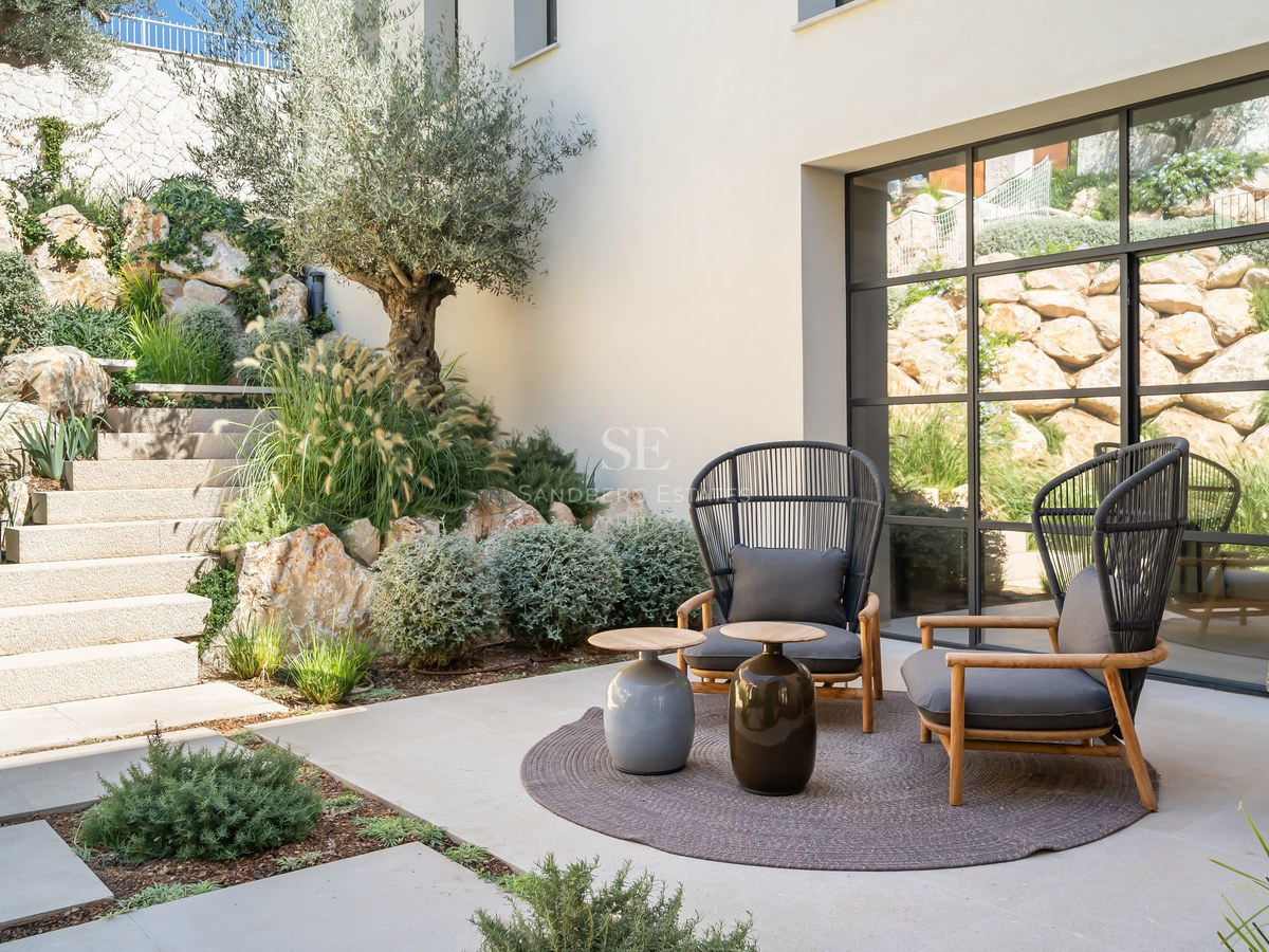 Modern patio with two grey armchairs, stone steps, olive tree, and large industrial-style window.