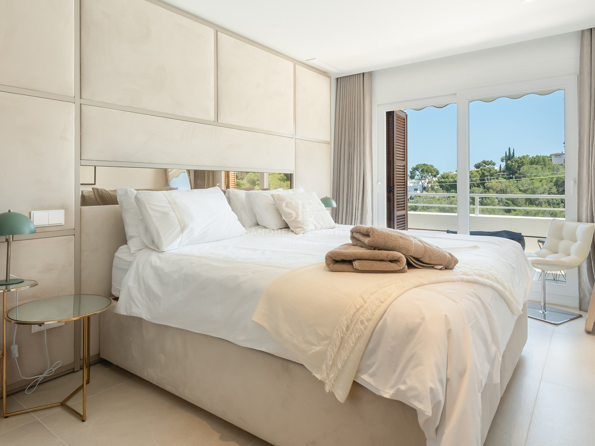 Ljust master bedroom med stoppad huvudgavel, glasbord och stora fönsterpartier mot trädgård.