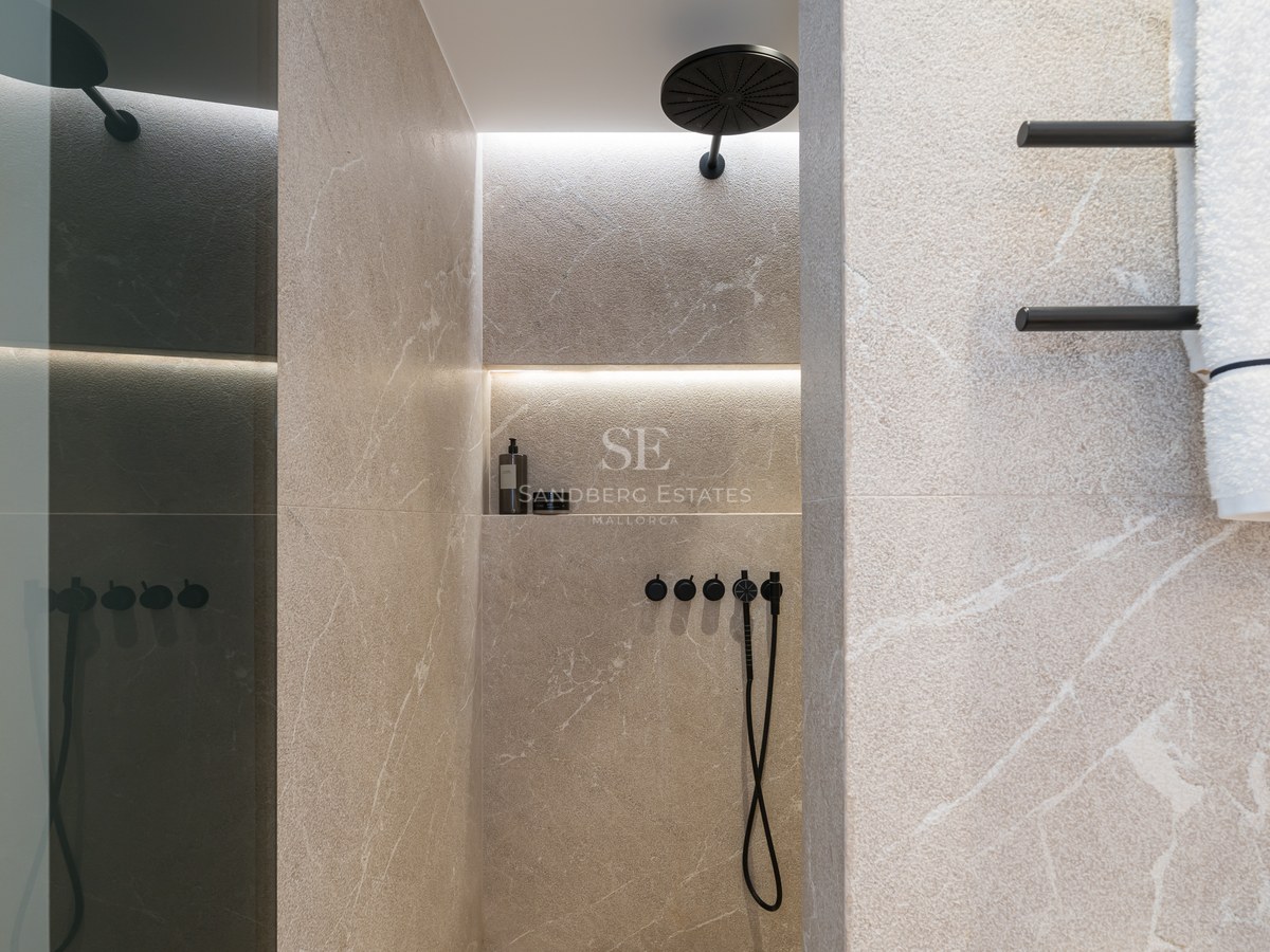 Douche avec carrelage en pierre beige, pommeau de pluie noir mat et niche éclairée sur un mur en pierre veinée.