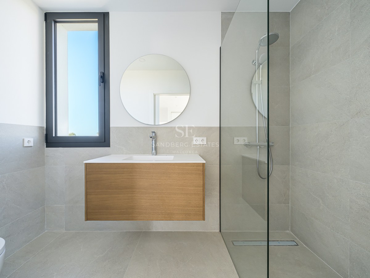 Salle de bain moderne avec carrelage gris, meuble en bois suspendu sous un miroir rond et douche à l'italienne vitrée.