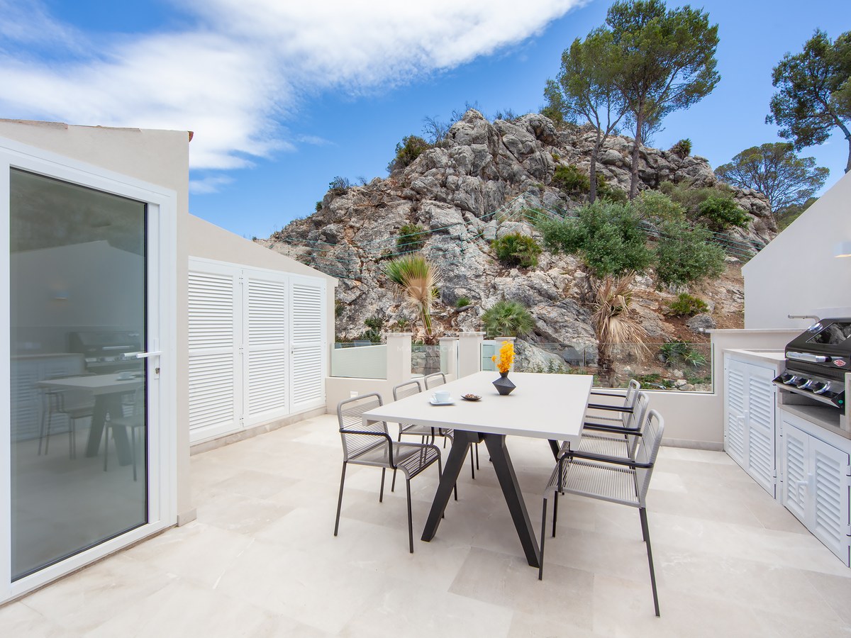 A modern white terrace with a dining table, chairs, and a barbecue area, set against a rocky mountain backdrop.
