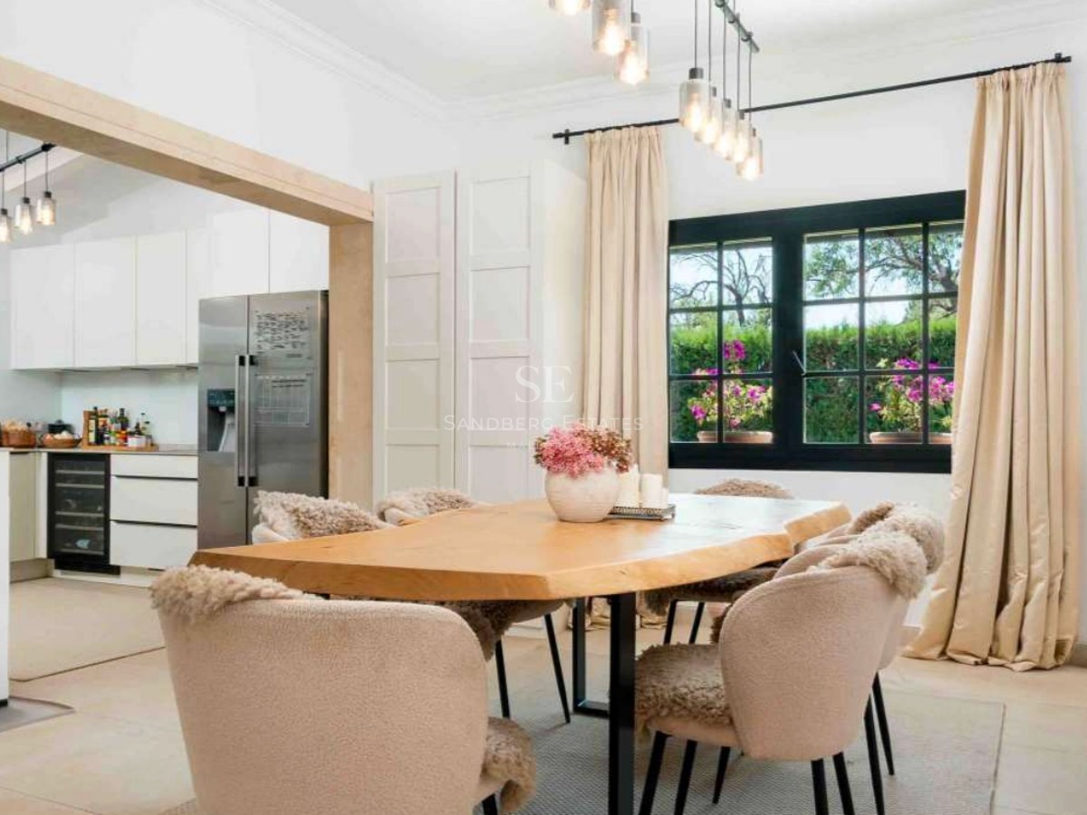Dining room featuring a live-edge wood table, beige chairs with fur accents, and a view into a modern white kitchen.