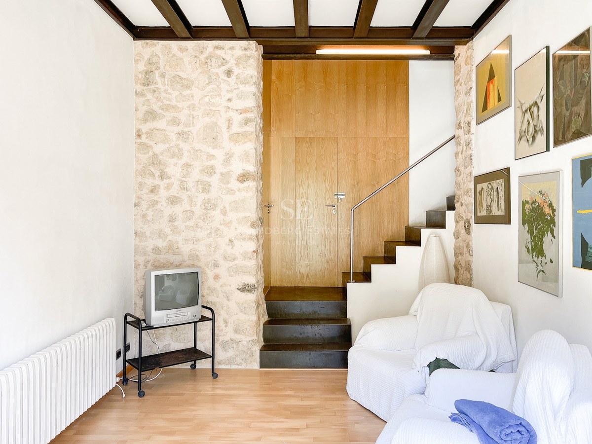 Salón con pared de piedra natural, vigas de madera oscura, escalera de pizarra y sillones blancos.