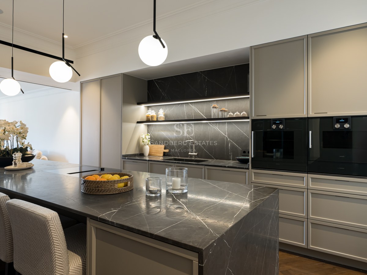 Cocina moderna con isla de mármol oscuro, armarios grises, electrodomésticos integrados y lámparas colgantes esféricas.