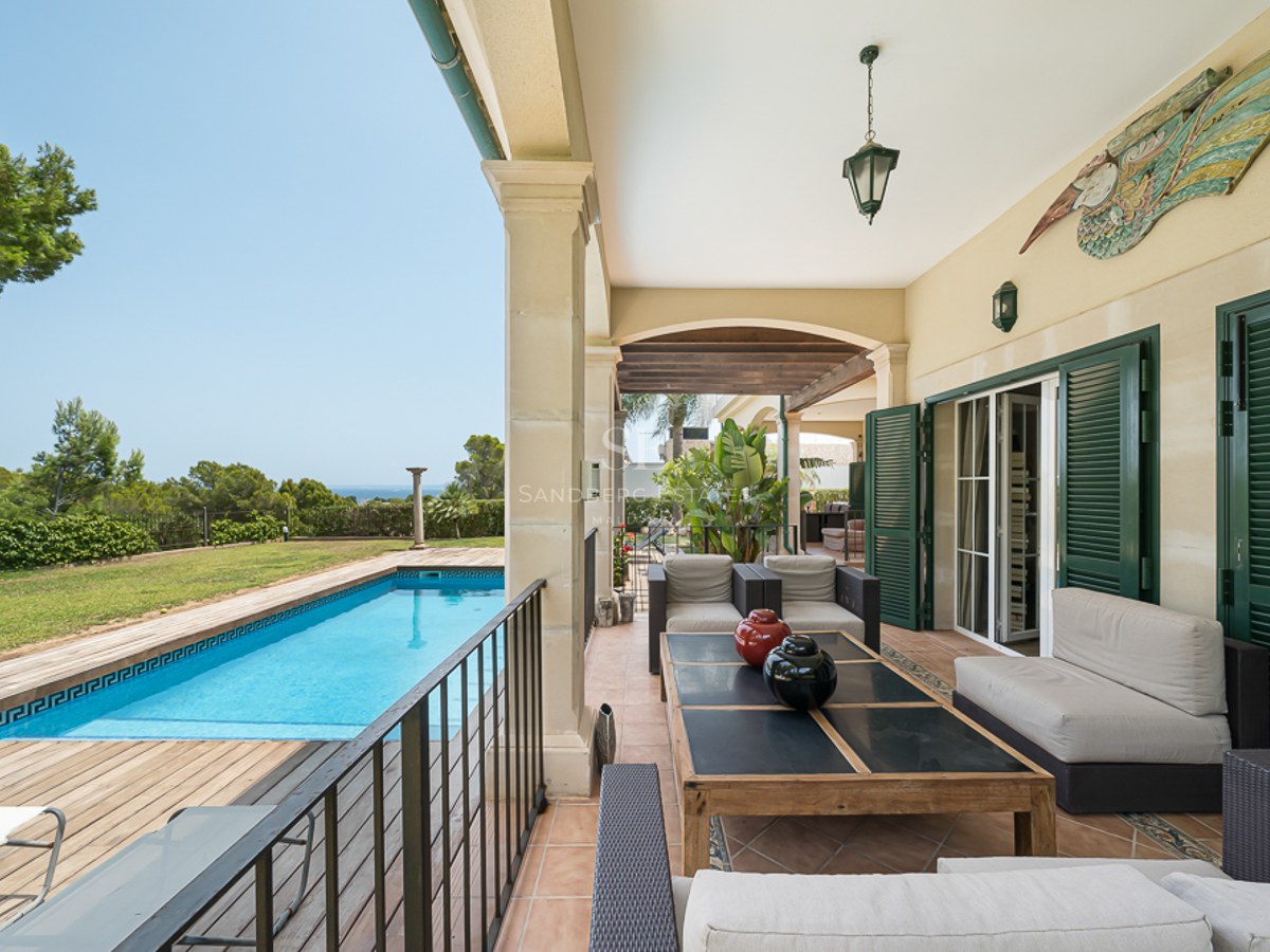 Porche couvert avec salon de jardin donnant sur une piscine, une terrasse en bois et la mer au loin.