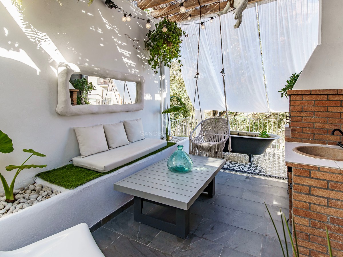 Terrasse blanche avec canapé intégré, balançoire macramé, évier en brique et baignoire noire sous des voiles.