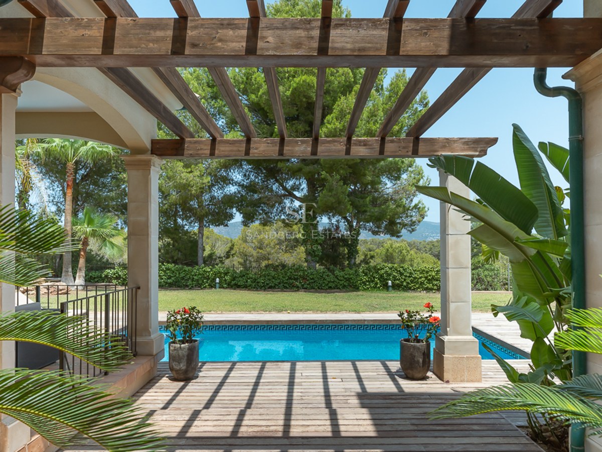 Vue d'une pergola en bois sur une piscine bleue entourée d'un jardin luxuriant et d'arbres méditerranéens.