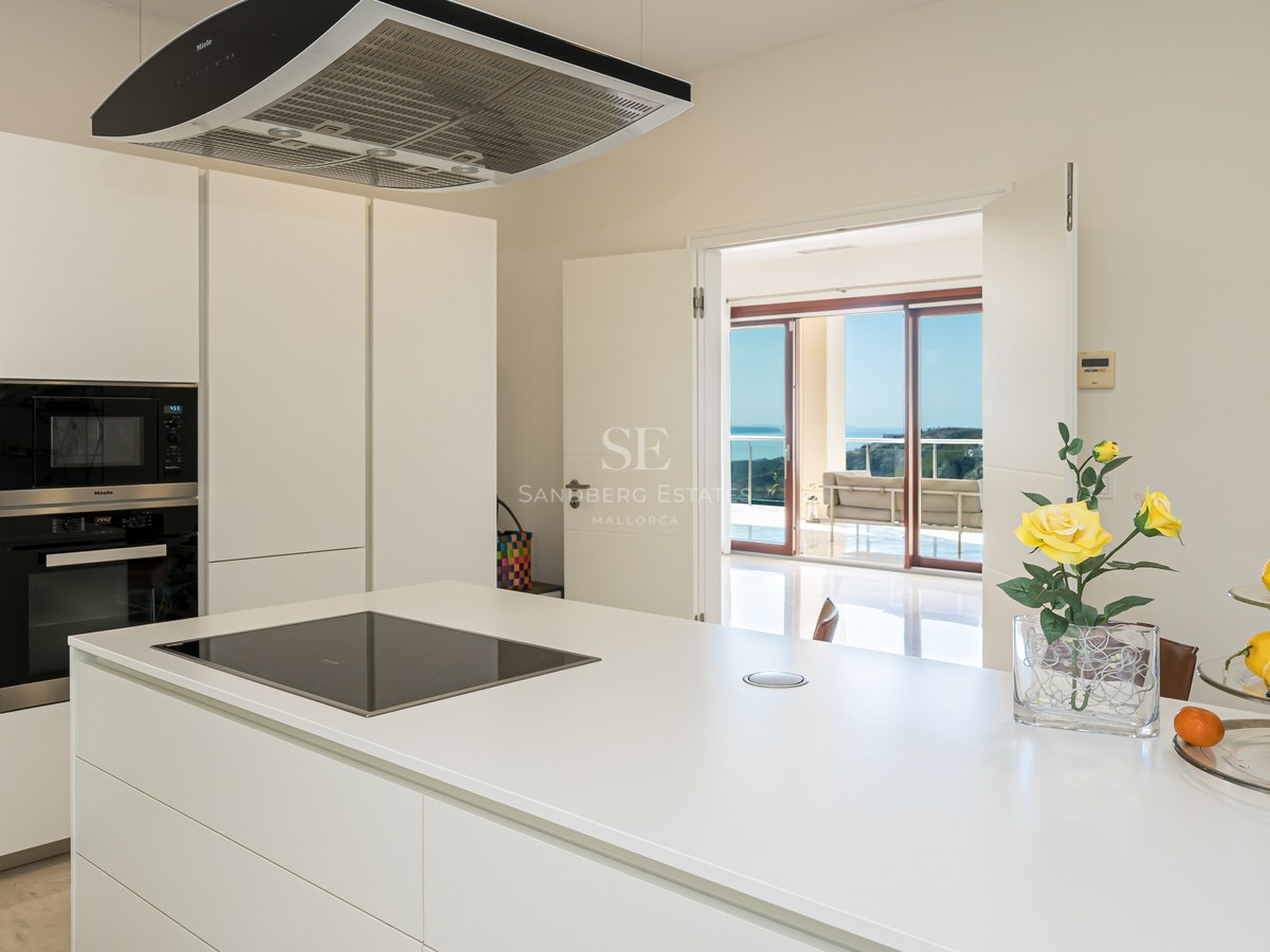 Cocina moderna blanca y luminosa con isla central, electrodomésticos Miele integrados y vistas a la terraza y el mar.