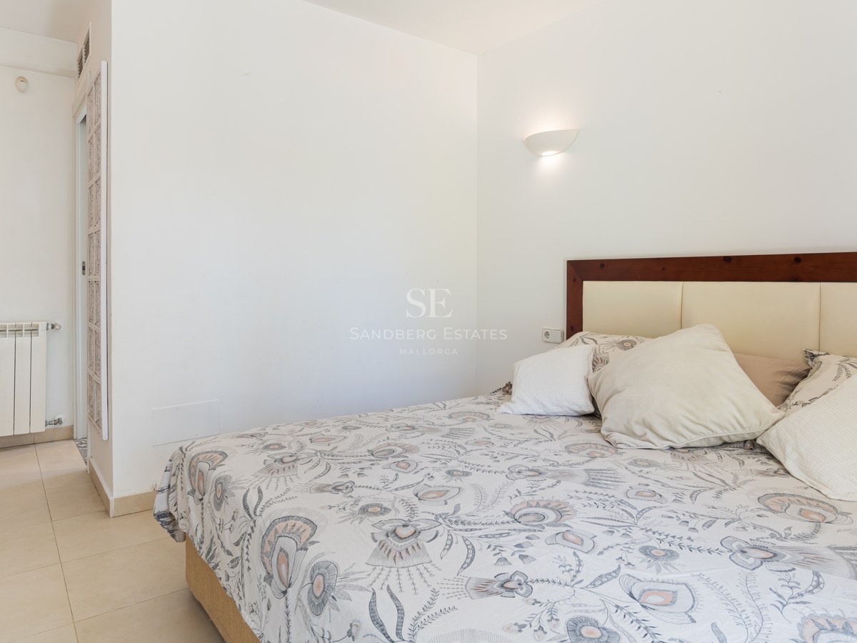 Dormitorio con sábanas estampadas, paredes blancas, suelo de gres crema y cabecero de madera con apliques.
