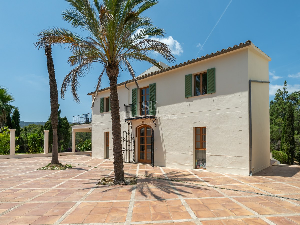 Two-story cream villa with green shutters and large terracotta courtyard with palm trees under a blue sky.
