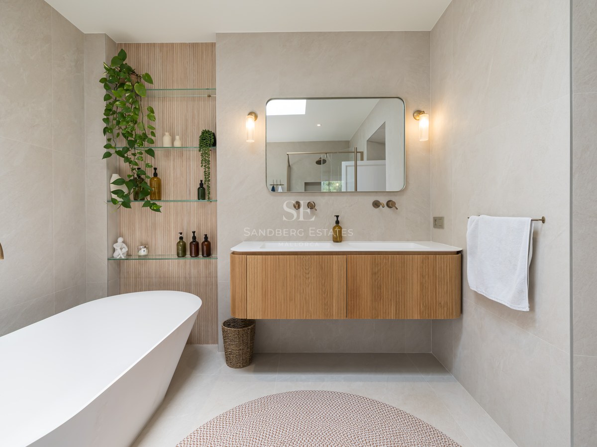 Contemporary bathroom featuring a white freestanding bathtub, floating fluted wood vanity, and brass fixtures against beige stone walls.