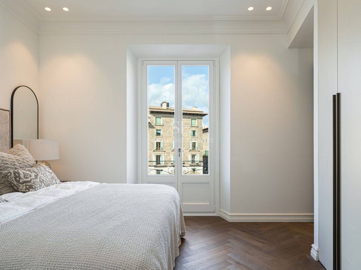 Dormitorio luminoso con suelo de espiga oscuro, armarios blancos y ventana a edificios históricos.