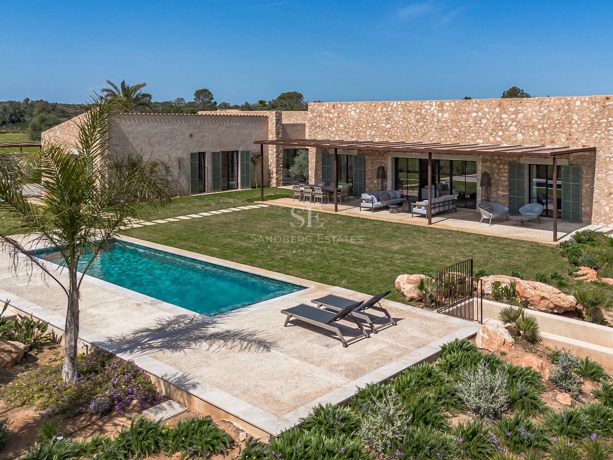 Vue extérieure d'une villa en pierre avec piscine, pelouse et terrasse ombragée sous un ciel bleu.