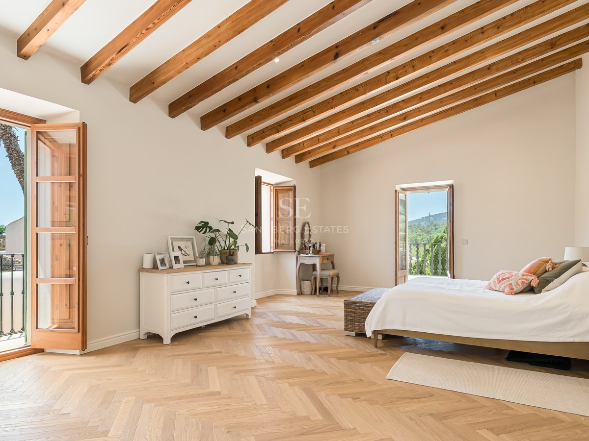 Spacious bedroom with high vaulted ceilings, wooden beams, herringbone oak floors, and a balcony overlooking a village.