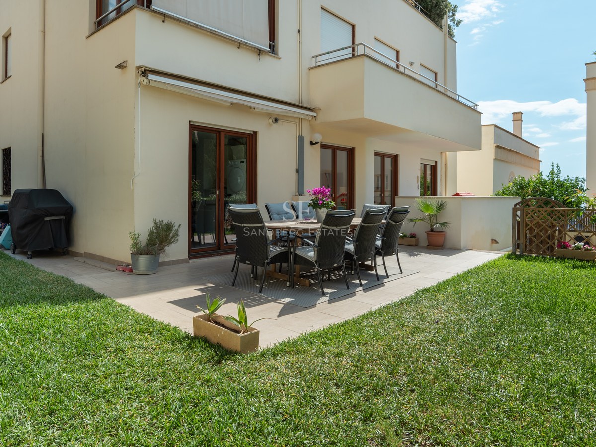 Patio exterior con mesa de comedor y sillas sobre terraza de baldosas rodeada de césped verde cuidado.