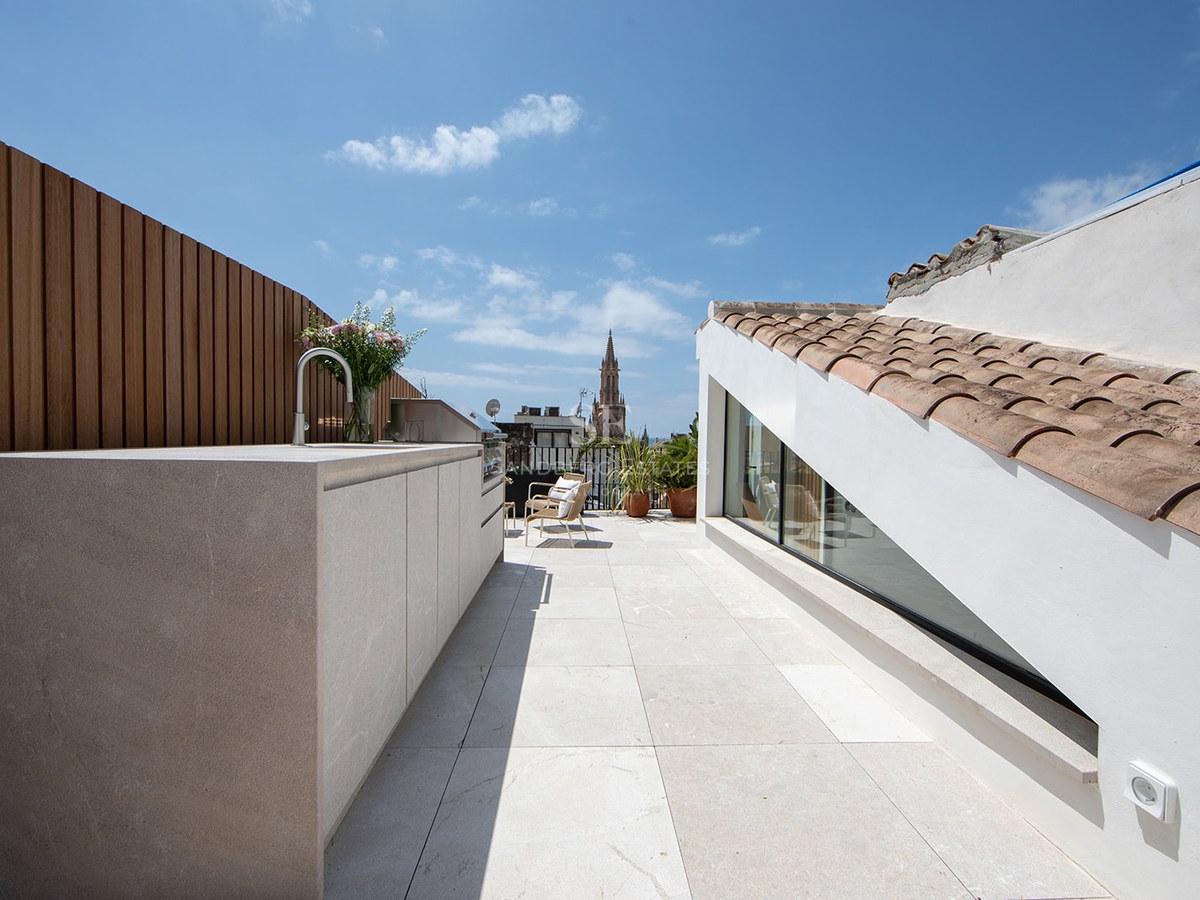 Modern rooftop terrace with outdoor kitchen, stone flooring, and a cathedral spire view under a blue sky.