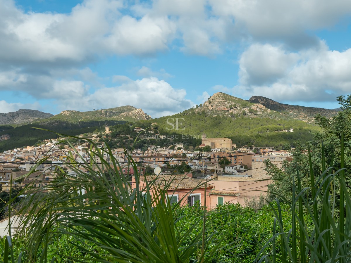 Vue d'un village méditerranéen aux toits de terre cuite entouré de montagnes verdoyantes sous un ciel nuageux.