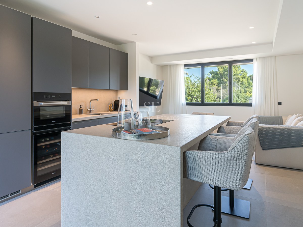 Cuisine grise moderne avec grand îlot en pierre, tabourets, appareils intégrés et vue sur la végétation.