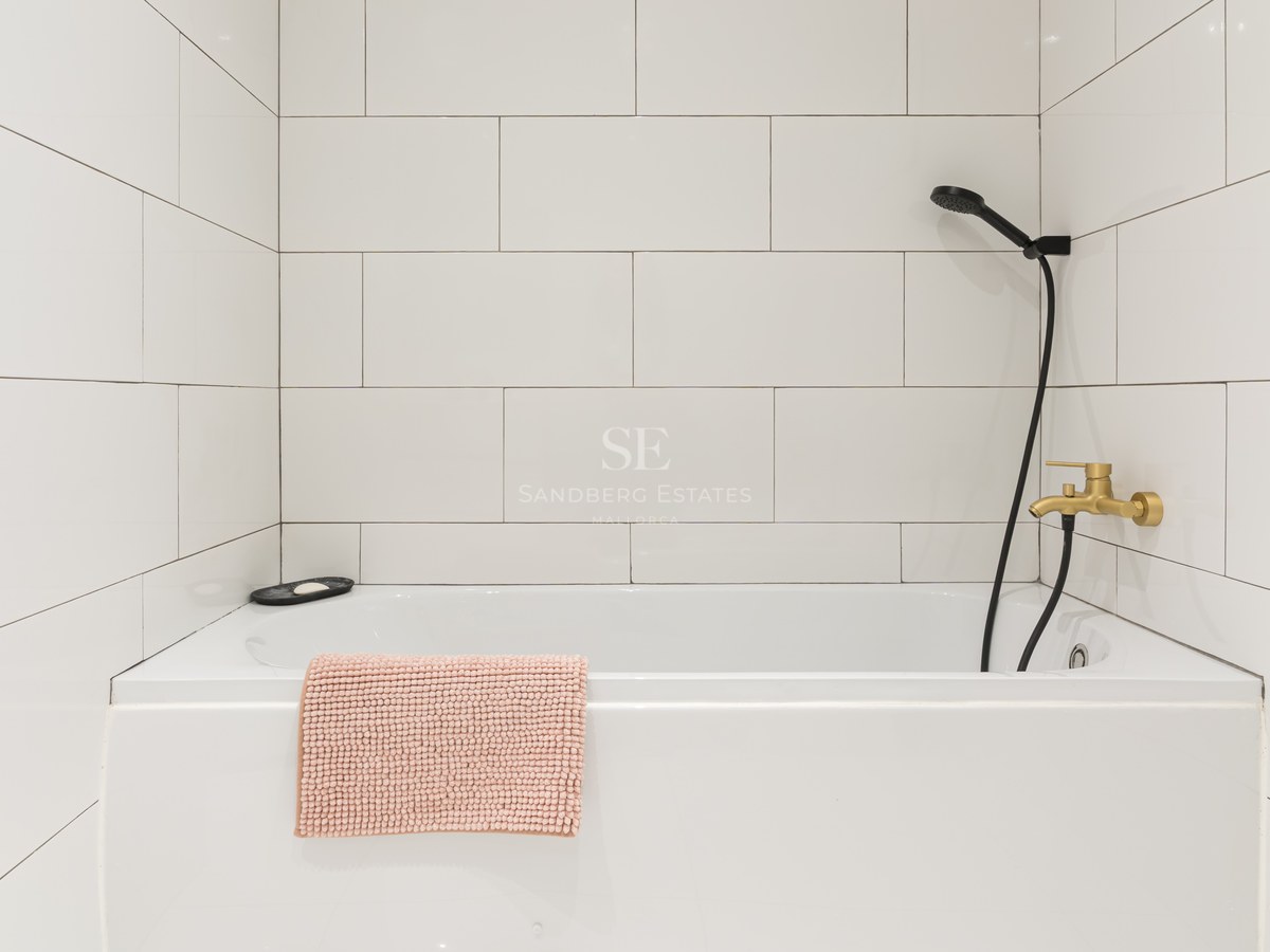 White bathtub with tiled walls, brass faucet, black shower head, and a textured pink bath mat.