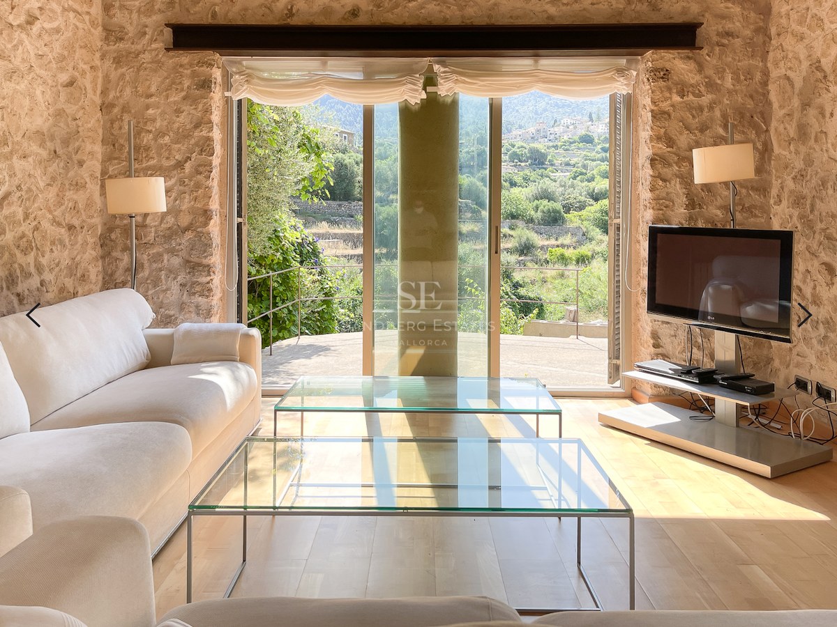 Salón luminoso con paredes de piedra natural, sofás blancos y grandes ventanales con vistas a montañas y vegetación.