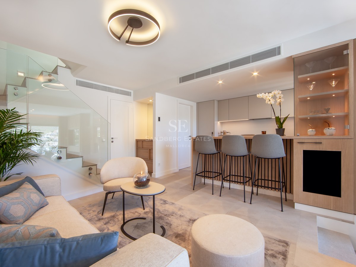 Modern living room with cream sofa, glass staircase, and a wood-paneled breakfast bar with grey stools.