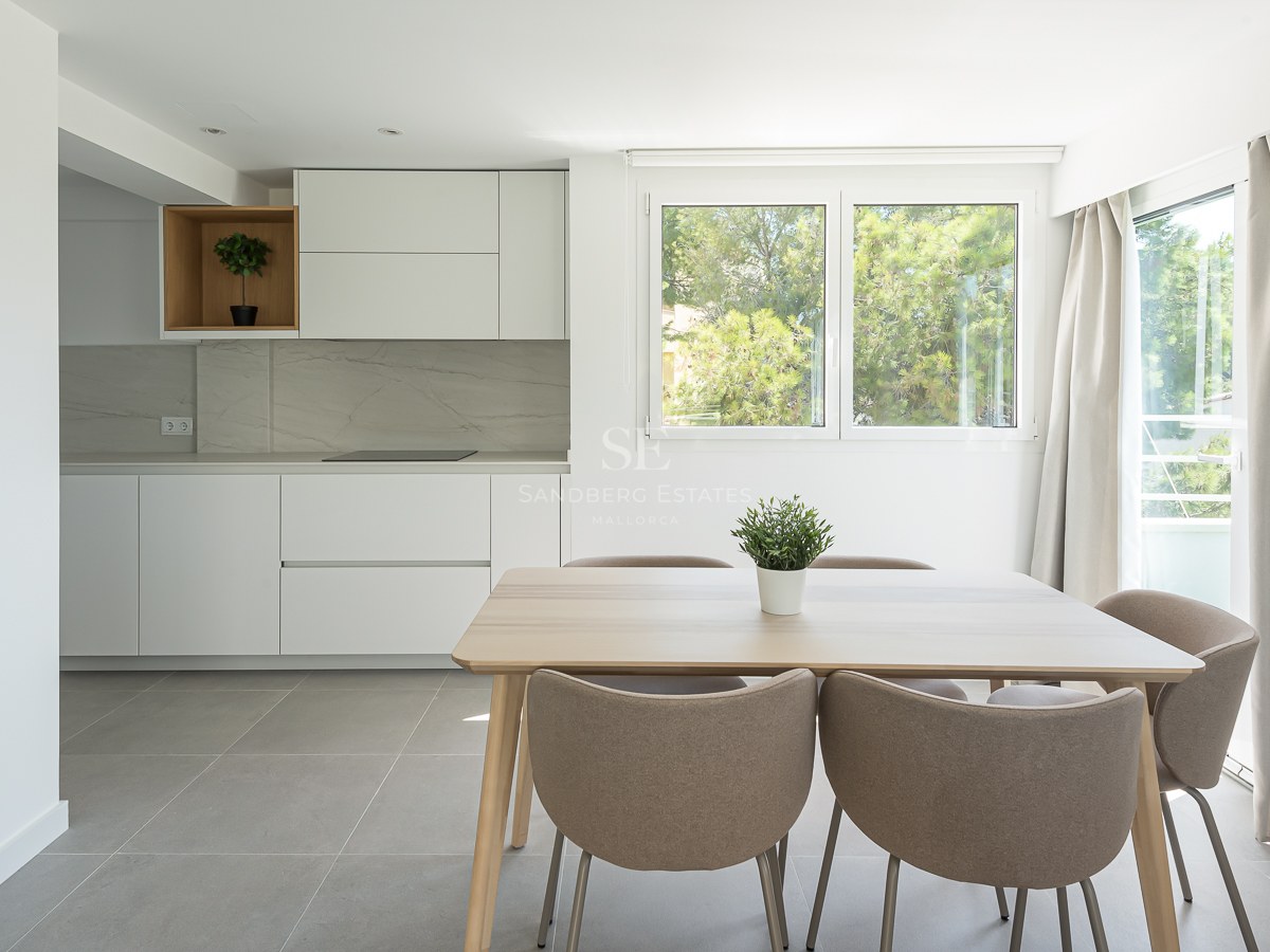 Salle à manger lumineuse avec table en bois clair, chaises taupe et cuisine blanche minimaliste vue sur jardin.