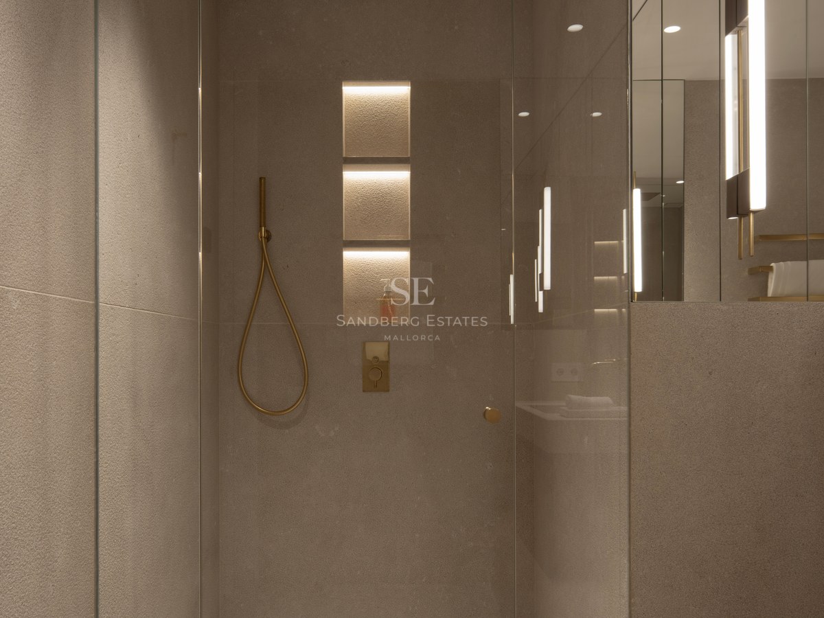 Douche en verre sans cadre avec carreaux de pierre couleur sable, éclairage encastré et robinetterie en or brossé.
