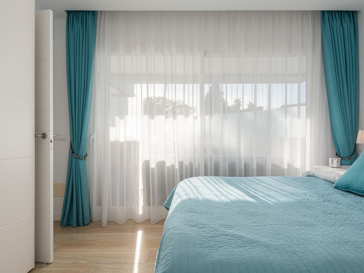 A bright bedroom with a teal bedspread and matching curtains, featuring floor-to-ceiling windows and wood flooring.
