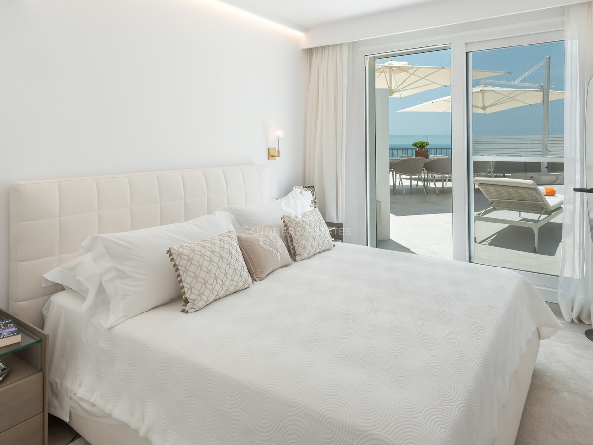 Bright master bedroom featuring a white tufted headboard and glass doors opening to a sun-drenched terrace with sea views.