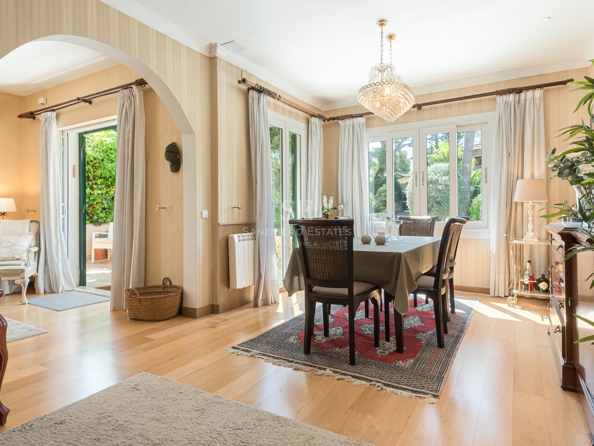 Salle à manger lumineuse avec lustre en cristal, meubles en bois sombre et portes-fenêtres ouvrant sur le jardin.