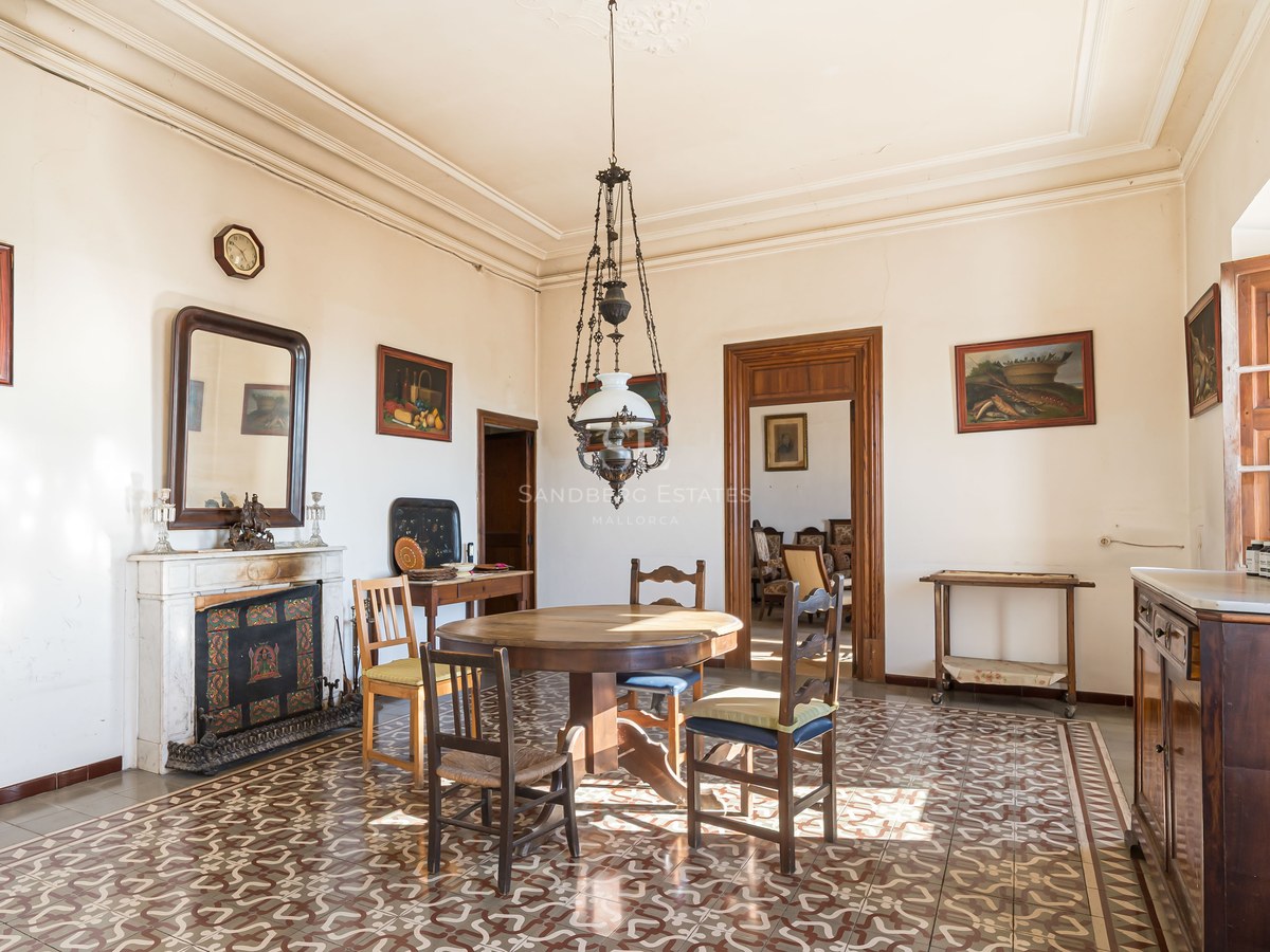 Comedor antiguo con mesa de madera, suelo de baldosas decoradas, chimenea de mármol y techos altos.