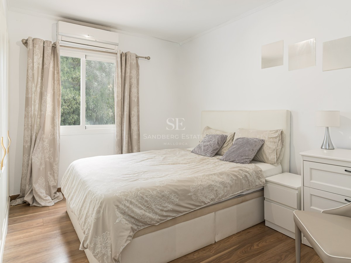 Bright bedroom with a double bed, white furniture, wooden floors, and a window showing green trees.