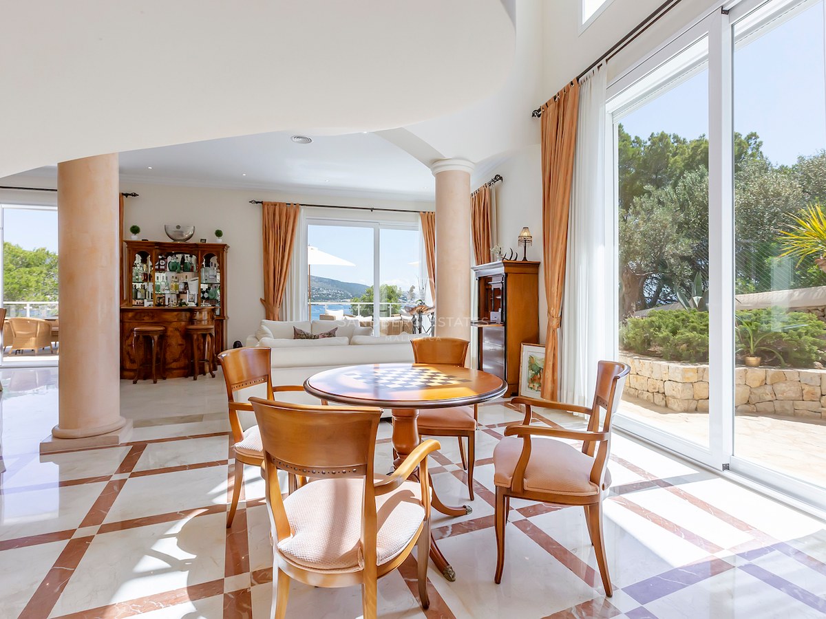 Spacious living area with marble floors, wooden table, pillars, and large windows overlooking a garden and sea.