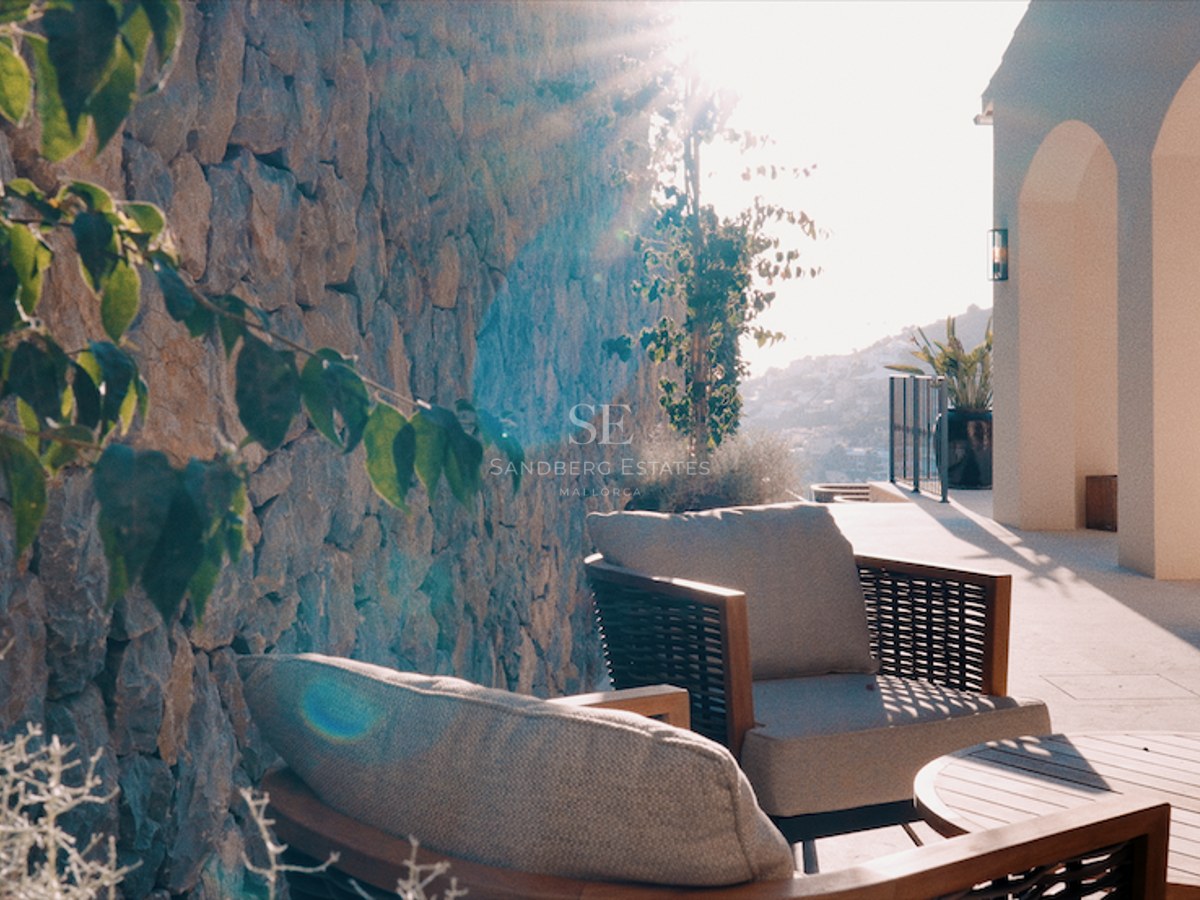 Sun-drenched terrace featuring elegant grey armchairs, a wooden table, a rustic stone wall, and white arched pillars.