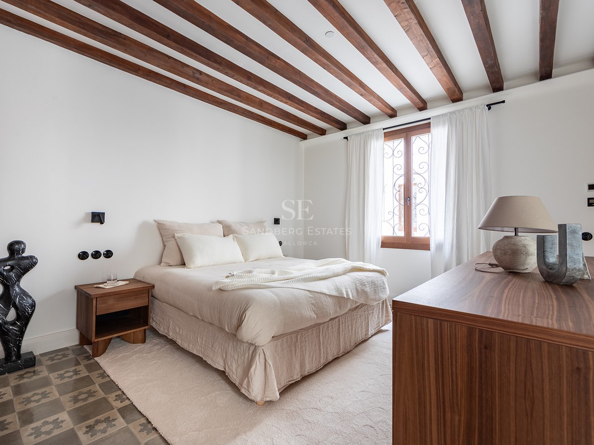 Bedroom featuring exposed dark wood ceiling beams, patterned floor tiles, and a window with sheer white curtains.