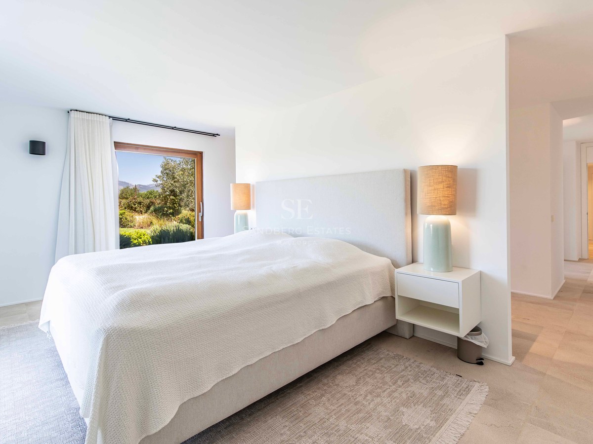 Chambre parentale lumineuse et moderne avec sol en pierre, lit rembourré et fenêtre donnant sur la verdure.