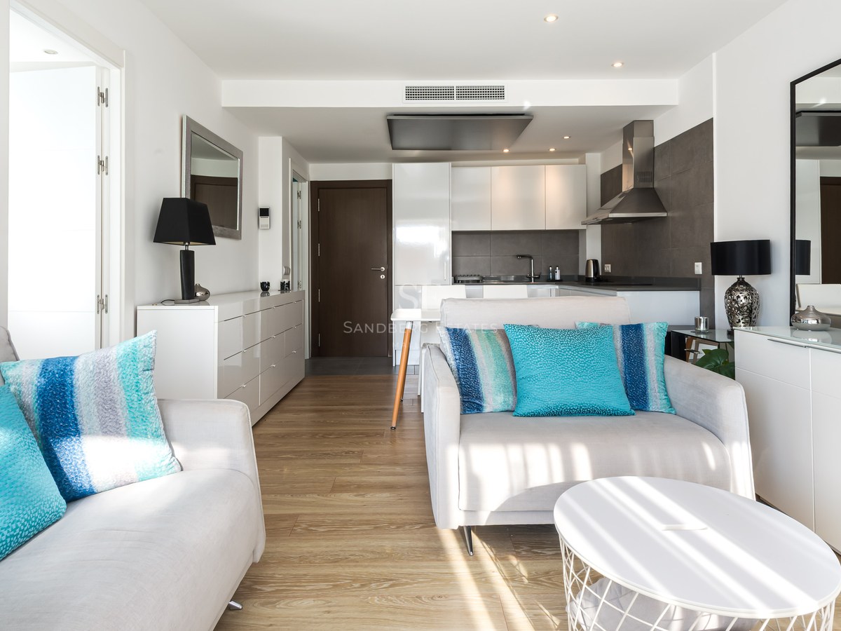 Bright living room with white sofas, turquoise cushions, wooden floors, and a modern kitchen in the background.