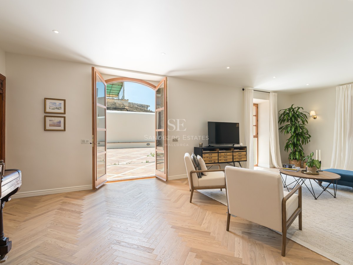 Bright living room featuring herringbone oak floors, cream armchairs, and glass doors opening to a sunlit terrace.
