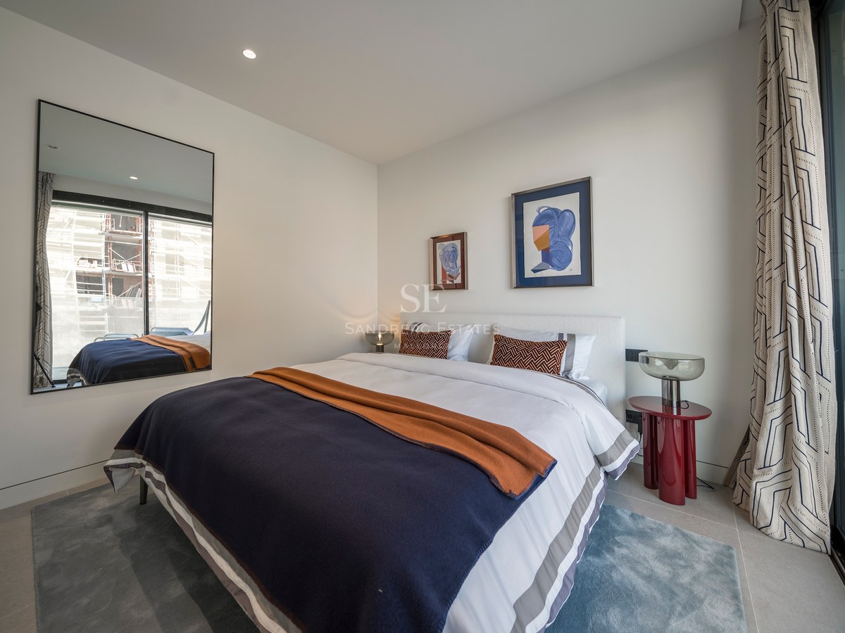 Chambre moderne lumineuse avec grand lit, literie bleu et orange, table rouge et un miroir reflétant une fenêtre.