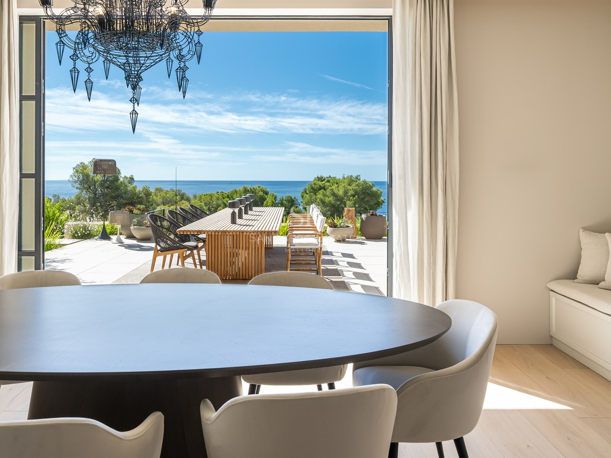Indoor dining area with dark wood table and chandelier opening onto a sunny terrace with Mediterranean sea views.
