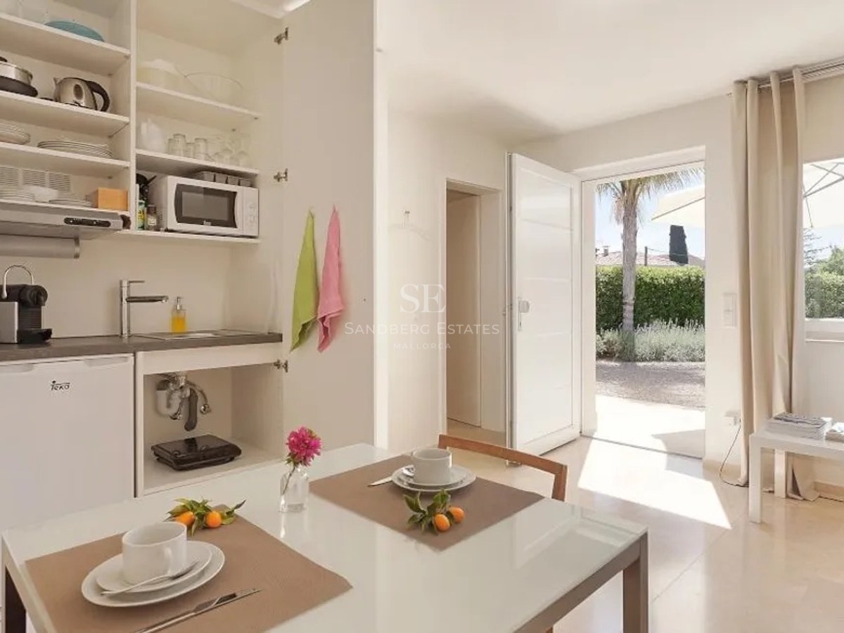 Modern white kitchenette and dining table with an open door looking out to a sunny garden and palm tree.
