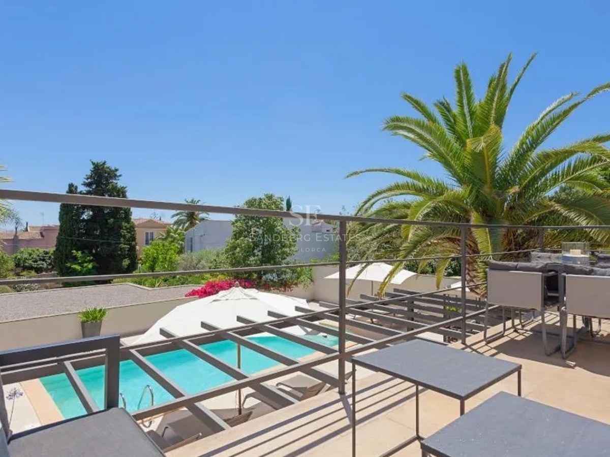 Elevated terrace with grey metal furniture and railings, overlooking a turquoise pool and lush palm trees under a blue sky.