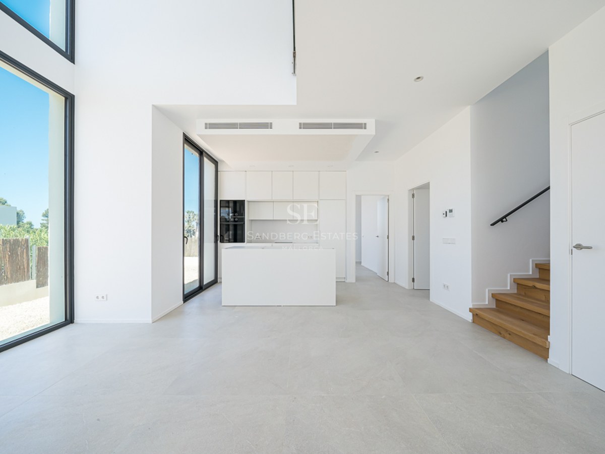 Cuisine blanche minimaliste avec îlot, escalier en bois et grandes baies vitrées du sol au plafond.