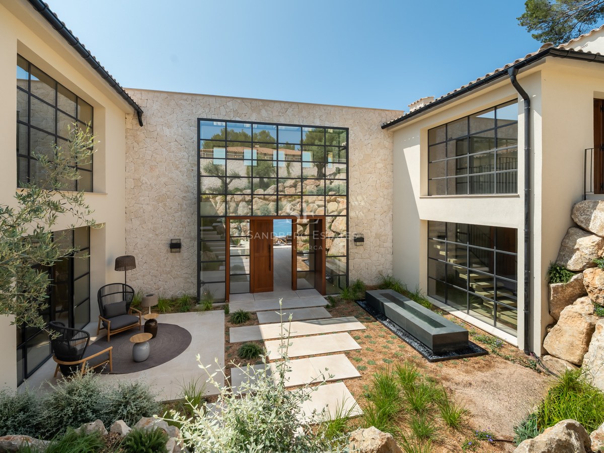 Modern villa entrance with a stone facade, large grid windows, and a landscaped courtyard with seating and water feature.