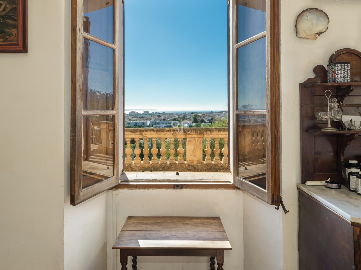 Ventana de madera abierta con vistas a una ciudad costera y al mar bajo un cielo azul despejado.